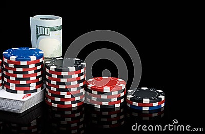 Stacks of poker chips stand on a deck of playing cards next to red cubes and a pack of hundred dollar bills on a glossy black Stock Photo