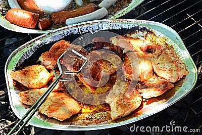Stainless steel barbecue tongs holding crunchy grilled chicken breast above alluminium plate, sausage and camembert plate in backg Stock Photo