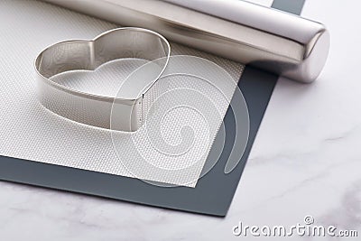 Stainless steel heart-shaped cookie cutter and rolling pin lying on baking mat Stock Photo