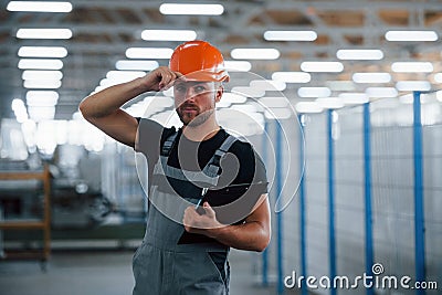 Stands in the corridor with documents in hands. Industrial worker indoors in factory. Young technician with orange hard hat Stock Photo