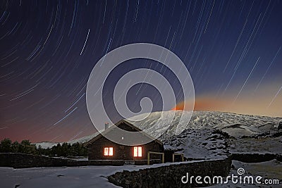Star trails on Galvarina Refuge with windows glowing under snowy Etna Volcano Stock Photo