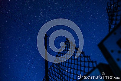 Starry sky with a silhouette of a pole with wires and a grid fence Stock Photo