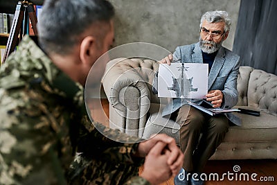 Start recovery. Mature psychologist holding picture with ink stain, Rorschach Inkblot in front of military man during Stock Photo