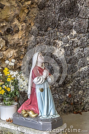 Statue of Bernadette praying to Our Lady of Lourdes with flowers next to her Stock Photo