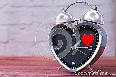 Steel alarm clock in the form of a heart, with a red heart on a pink wooden table and with a white brick wall. Valentine`s Day Stock Photo