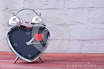 Steel alarm clock in the form of a heart, with a red heart on a pink wooden table and with a white brick wall. Valentine`s Day Stock Photo