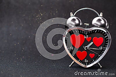 Steel clock alarm in the form of a heart with red hearts inside on a black glitter shiny background. Happy Valentine`s Day Stock Photo