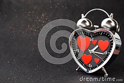 Steel clock alarm in the form of a heart with red hearts inside on a black glitter shiny background. Happy Valentine`s Day Stock Photo