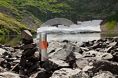 A steel thermos stands on the shore of a mountain lake on a summer day. Stock Photo