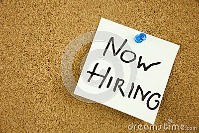 A sticky note on a cork board with the words now hiring on it Stock Photo
