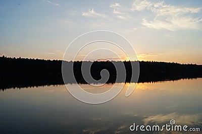 A still dark moody lake at dusk reflects the setting sun on the water surface with a silhouette of trees Stock Photo