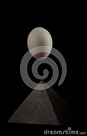 Still-life. A large white egg hangs above a black pyramid on a black background with a reflection Stock Photo