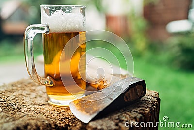 Still lifephoto of glass of fresh cold beer on wooden stub alongside axe. Rural concept, masculinity, coarse, rough wilderness Stock Photo