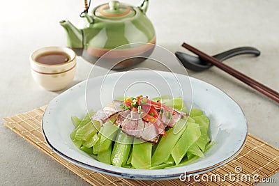 Stir-fried Chinese Lettuce with Sichuan Smoked Bacon Bits in a dish isolated on wooden mat side view on grey background Stock Photo