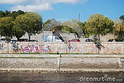 Stone fence with grafitti by river Nisava and cars on the street with trees under blue sky Editorial Stock Photo