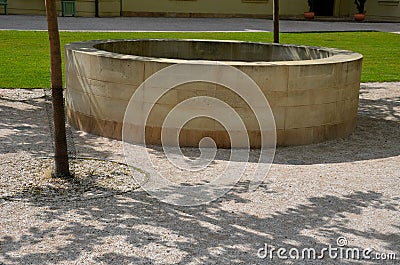 Stone round fountain with a protective grid of twisted prisms. forged steel bars go spiraled at the top. around the well there is Stock Photo