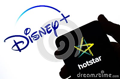 Hotstar logo on the silhouette of a smartphone hold in hand and Disney+ logo at the background. Concept. Real photo, not a montage Editorial Stock Photo