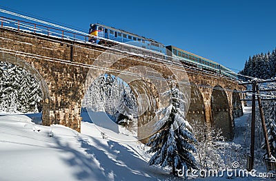 Stone viaduct arch bridge on railway through mountain snowy fir forest and locomotive with a passenger train. Snow drifts on Stock Photo