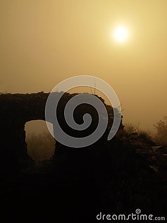 The stony ruin of medieval stronghold tower on hill. Early morning sunshine hidden in heavy mist. Stock Photo