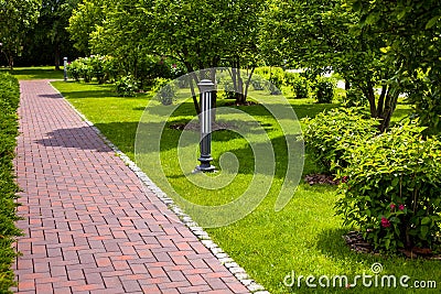 Straight stone tile pavement in a park landscaped with green grass and bushes. Stock Photo