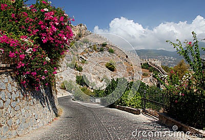 A street with bright pink flowers and a view of part of the Alanya Kalesi fortress in Alanya, Turkey Stock Photo