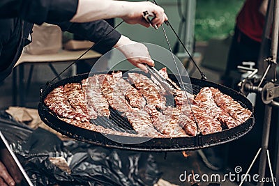 street food festival. grilled ribs meat. man holding steel tongues and roasting beef pork on big grill. chef in black form Stock Photo