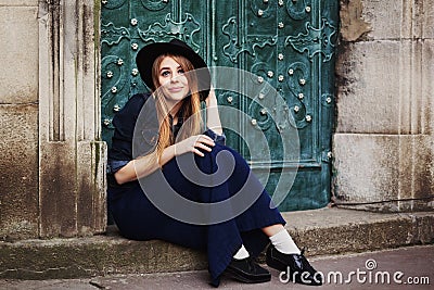 Street full body portait of stilylish smiling young woman sitting near the door. Model looking at camera. City lifestile Stock Photo