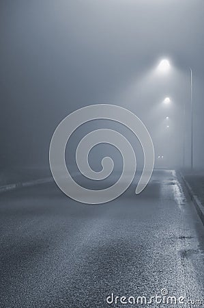 Street lights, foggy misty night, lamp post lanterns, deserted road in mist fog, wet asphalt tarmac, car headlights approaching Stock Photo