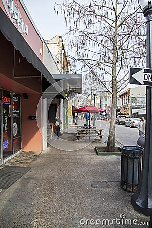 A street lined with shops and restaurants with tall black light posts and cars on the street and people on the sidewalk Editorial Stock Photo
