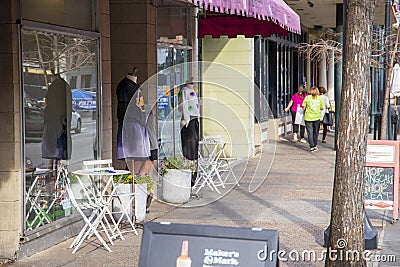 A street lined with shops and restaurants with tall black light posts and cars on the street and people on the sidewalk Editorial Stock Photo