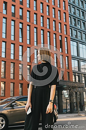 Street portrait of a stylish girl in dark casual clothes stands on the street of a metropolis and a modern building, looking away Stock Photo