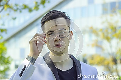 Street portrait of a thoughtful young man 18-20 years old in a white jacket and glasses on the background of a business center, sm Stock Photo