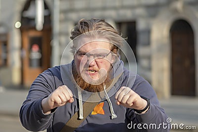 Street portrait of a 25-30-year-old man with glasses with an angry expression, wants to fight, on a city background, medium plan. Stock Photo