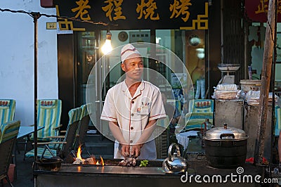 Street scene in the city of Dunhuang, with a cook preparing lamb kebab at a food stall, in China Editorial Stock Photo