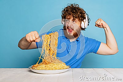 Emotive young hairy red-bearded man tasting large portion of noodles, pasta isolated on blue studio background. Stock Photo