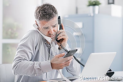 Stressed desperate businessman working in his office and having multiple calls, he is holding two handsets and a mobile phone, Stock Photo