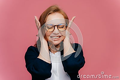 Stressed female teacher covers ears, has headache because of noisy pupils, clenches teeth, ignores noise, wears optical glasses, Stock Photo