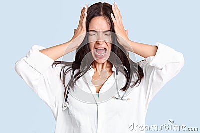 Stressful female doctor or specialist holds hands on head, suffers from terrible headache, exlaims from pain, keeps mouth widely o Stock Photo