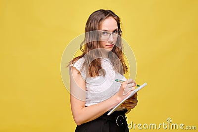 Strict teacher girl with glasses on a yellow background makes a note with a pen in a notebook Stock Photo