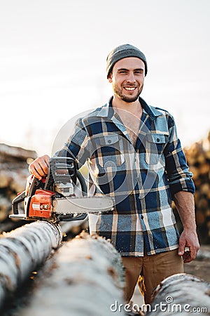 Strong bearded woodcutter wearing plaid shirt hold chainsaw in hand on background of sawmill and warehouse of trees Stock Photo