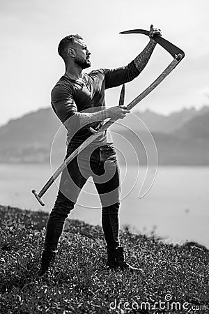 Strong muscular power sexy farmer using scythe. Farmer with a scythe on green grass field. Guy cut grass. Rural farmer Stock Photo