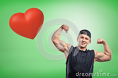 Strong muscular young man showing biceps with big red heart on green background Stock Photo