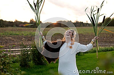 Strong young woman with a bulrush grass reed on rural field background at sunset Stock Photo