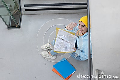 A student in a blue sweatshirt and yellow hat sits with folders between the stairs and greets a friend Stock Photo