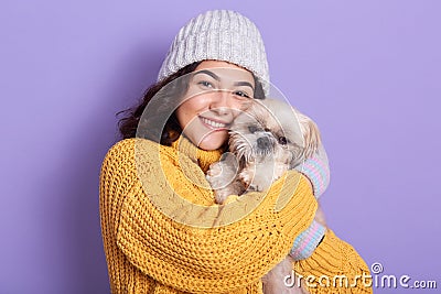 Studio portrait of cute young woman wearing cap and yellow sweater, looks smiling at camera and hugging her puppy. Girl playing Stock Photo