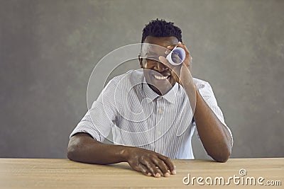 Happy black man sitting at desk and looking through hole in paper roll like in telescope Stock Photo