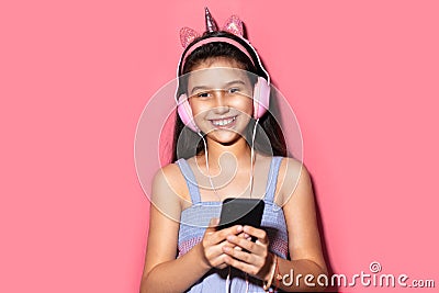 Studio portrait of happy little child girl, holding smartphone in hands, using headphones. Background of pastel pink color. Stock Photo