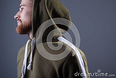 Studio portrait of a young bearded guy of twenty-five years old. in a sports jacket with a swamp-colored hood. On a gray Stock Photo