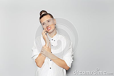 Studio portrait of young happy disappoint girl with hair bun talking on smartphone, on grey background, looking up, wearing white Stock Photo