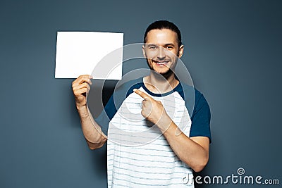 Studio portrait of young happy man holding white empty paper board with pointed finger, on background of blue color Stock Photo